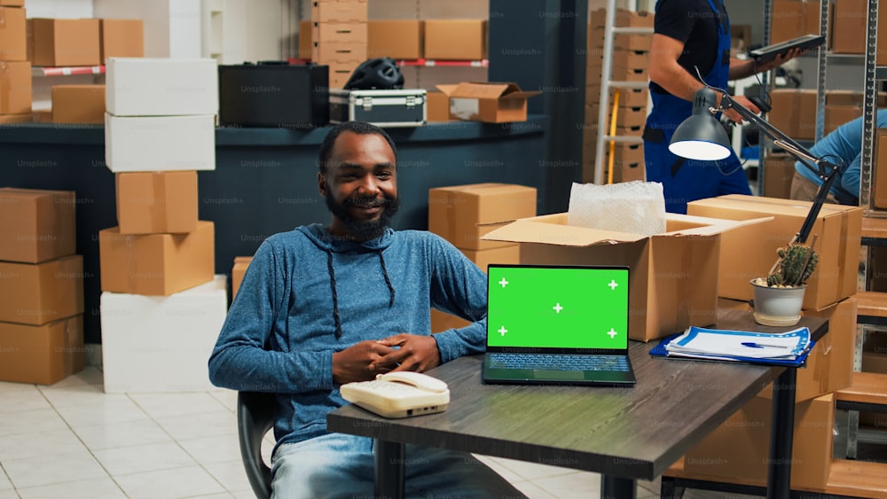 Homem afro-americano usando laptop com modelo de tela verde, planejando o envio de mercadorias no armazém. Funcionário do sexo masculino trabalhando com display isolado e tela de copyspace cromakey.