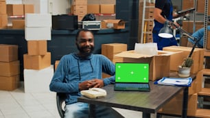 African american man using laptop with greenscreen template, planning merchandise shipment in warehouse. Male employee working with isolated display and chromakey copyspace screen.