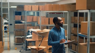 Male entrepreneur taking inventory notes on clipboard files, checking quality of goods or products and packages. Young employee looking at cardboard boxes, merchandise stock logistics.