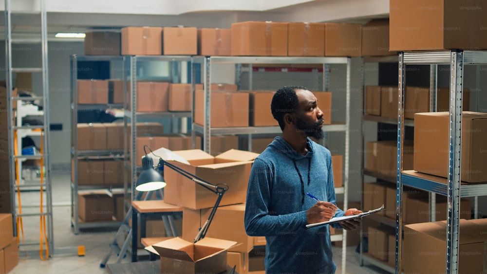 Male entrepreneur taking inventory notes on clipboard files, checking quality of goods or products and packages. Young employee looking at cardboard boxes, merchandise stock logistics.