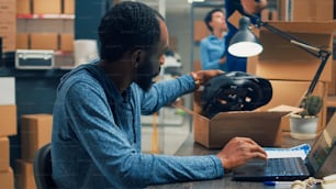 African american worker analyzing quality of merchandise in warehouse, preparing products order for shipment. Young man putting supplies and goods in carton boxes to ship to customers.