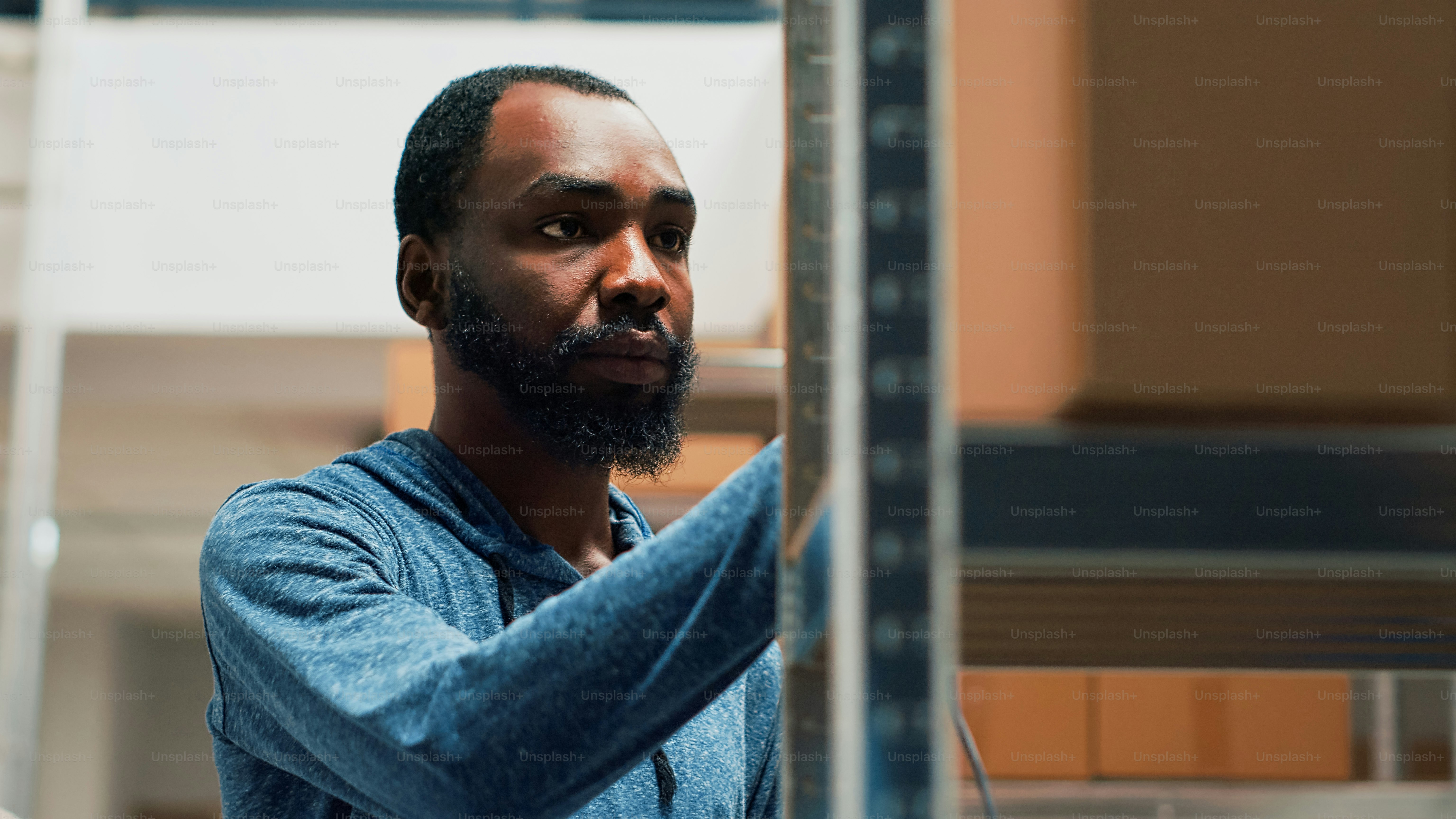 Afroamerikanischer Arbeiter, der Scanner und Tablet verwendet, um den Lagerbestand zu überprüfen und an der Produktverwaltung zu arbeiten. Junger Mann scannt Waren in Pappkartons in Lagerregalen. Aufnahme aus der Hand.