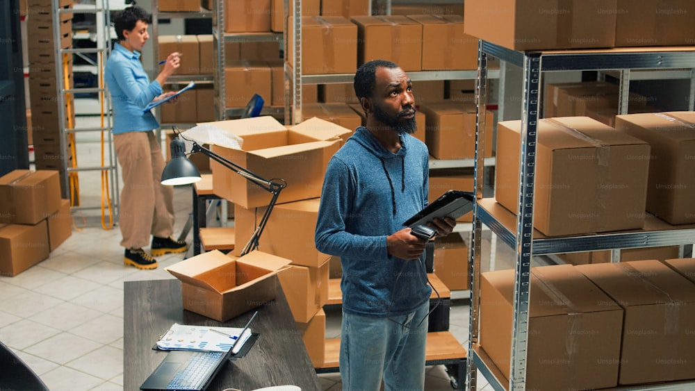 Small business owner scanning bar code on carton boxes, working on doing merchandise inventory in warehouse space. Male worker taking packages from sorage room racks and checking them.