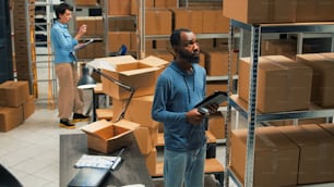 Small business owner scanning bar code on carton boxes, working on doing merchandise inventory in warehouse space. Male worker taking packages from sorage room racks and checking them.