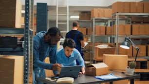 Offce employees doing teamwork to ship goods in cardboard boxes, checking quality of merchandise in storehouse space. Diverse owners using laptop to plan products shipment distribution.