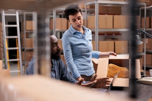 倉庫のラップトップコンピュータでクライアントの出荷データをチェックしながら、従業員に流行の服の入ったパッケージを示すカートンボックスを持つ倉庫監督者。配送センターフルフィルメント会社
