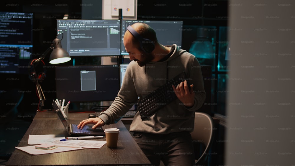 IT engineer having fun playing air guitar at night, using keyboard as instrument and fooling around. Male programmer enjoying music on headset, coding new database server. Handheld shot.