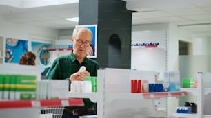 Senior man analyzing boxes of medicine and pills bottles, looking at pharmaceutical products to buy prescription treatment. Buying drugs and medicaments to cure disease at pharmacy shop.