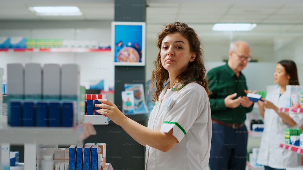 Portrait of pharmacist working at drugstore counter with medicaments and boxes of vitamins on shelves. Medical employee at pharmacy desk to sell supplements and medicine products. Handheld shot.