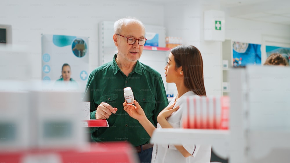 Asian pharmacist showing medicaments boxes to senior client, explaining disease treatment and supplements in pharmacy store. Medical worker talking to eldery man about pharmaceutics. Handheld shot.