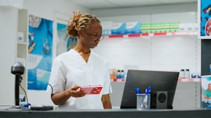 Happy african american woman sitting at pharmacy desk and helping clients with prescription medicine, holding pills bottles and boxes of vitamins. Working at drugstore counter. Handheld shot.