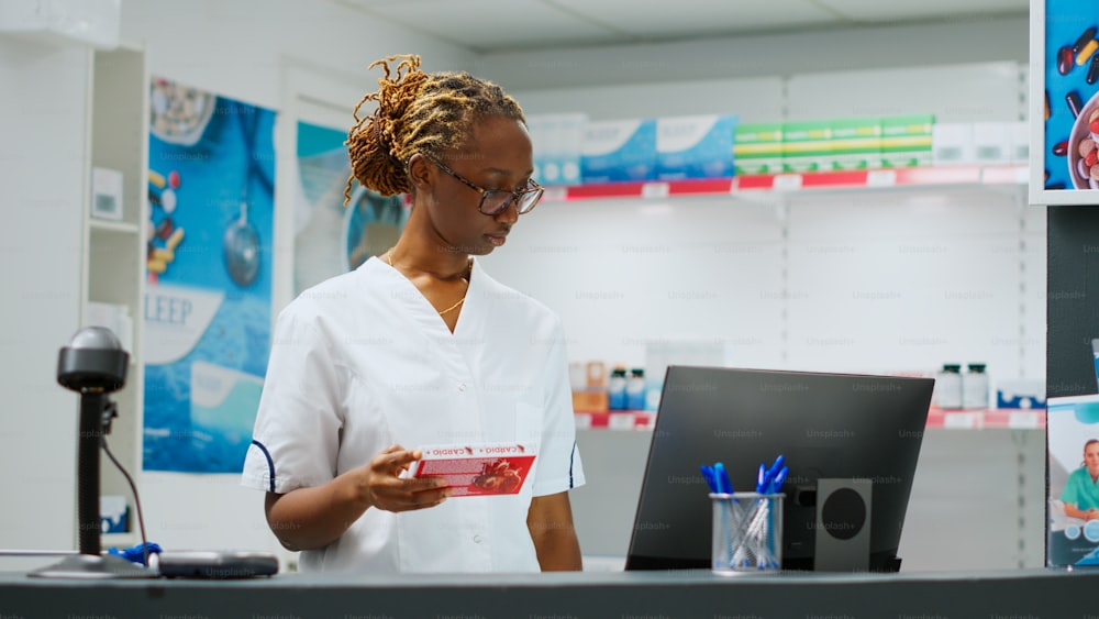 Happy african american woman sitting at pharmacy desk and helping clients with prescription medicine, holding pills bottles and boxes of vitamins. Working at drugstore counter. Handheld shot.