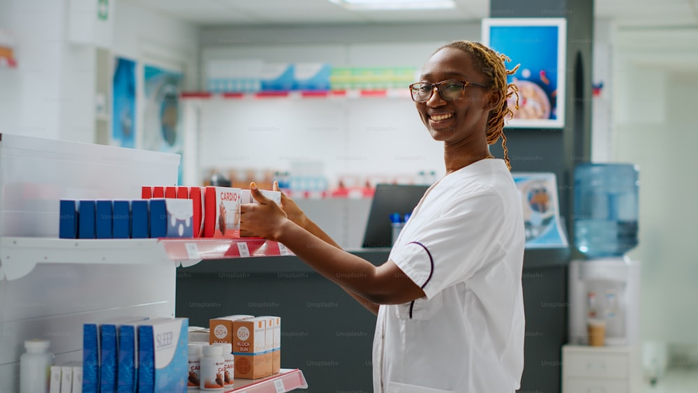 Lächelnder afroamerikanischer Apotheker, der Medikamente in den Regalen der Apotheke überprüft, Kunden mit Medikamenten hilft, Pillenflaschen und Schachteln mit Vitaminen hält. Arbeiten an der Drogerietheke. Aufnahme aus der Hand.