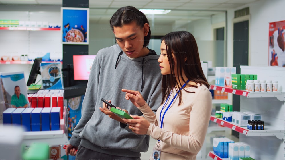 Female pharmacist looking at smartphone to give pills to young man, searching for medical treatment on shelves. Showing drugs and pharmaceutical products, pharmacy healthcare medicaments.