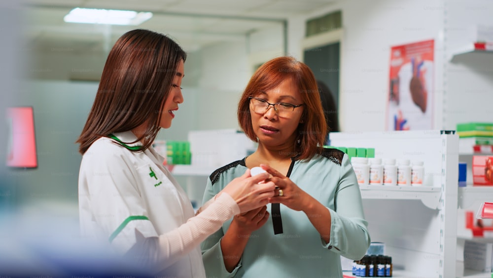 Medical consultant showing cardiology pills to asian woman in pharmacy, explaining treatment to cure heart problems. Female pharmacist talking to client about medical products. Handheld shot.