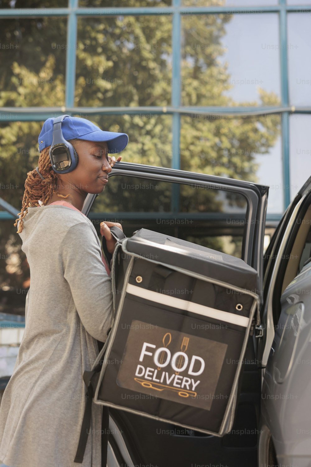 Young courier in headphones delivering restaurant food order by car. Delivery service worker listening to music in earphones, taking out backpack from automobile, medium shot