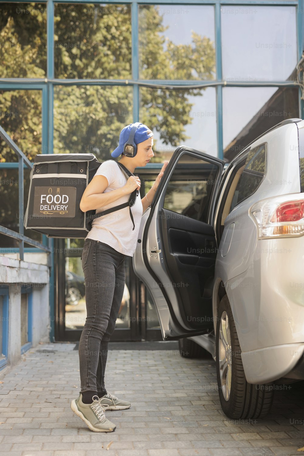 Office lunch delivery service courier delivering food by car, holding backpack with food. Woman listening to music in headphones, carrying bag with meal, standing near vehicle, side view