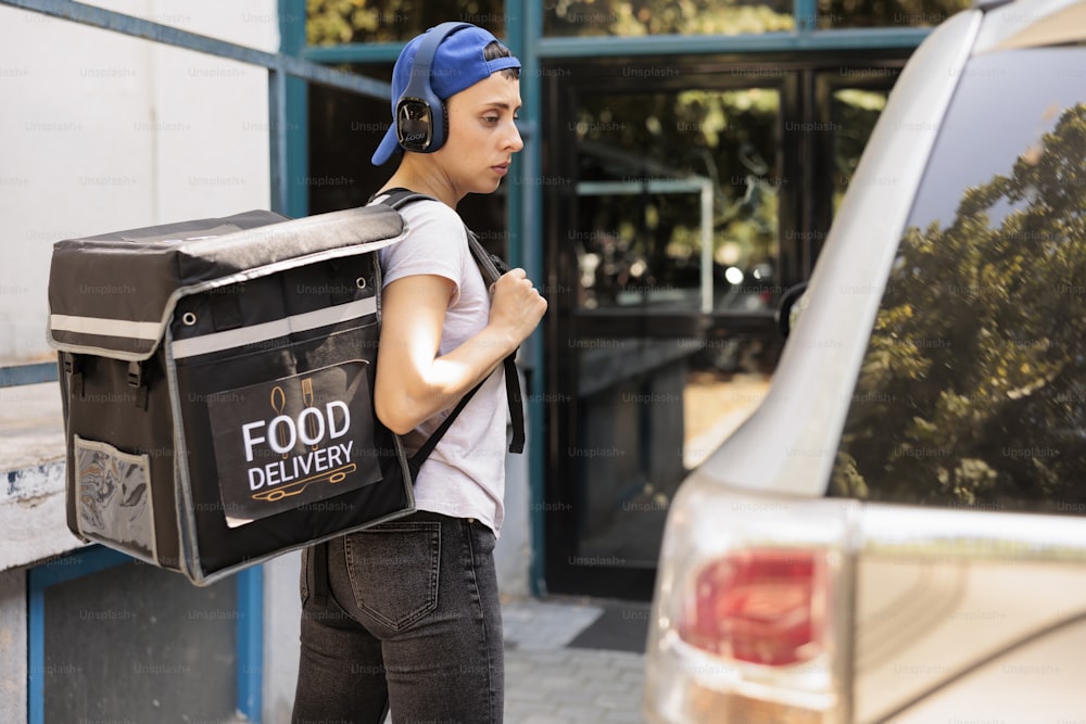 車で食品注文を配達するサーマルバッグ付きの宅配便、昼食付きのバックパックを運ぶ。オフィス給食サービス ヘッドフォンで音楽を聴く若い従業員、側面図