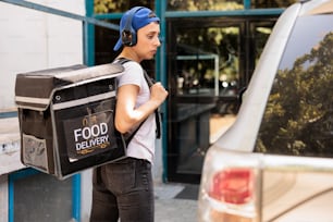 Courier with thermal bag delivering food order by car, carrying backpack with lunch. Office meal delivery service young employee listening to music in headphones, side view