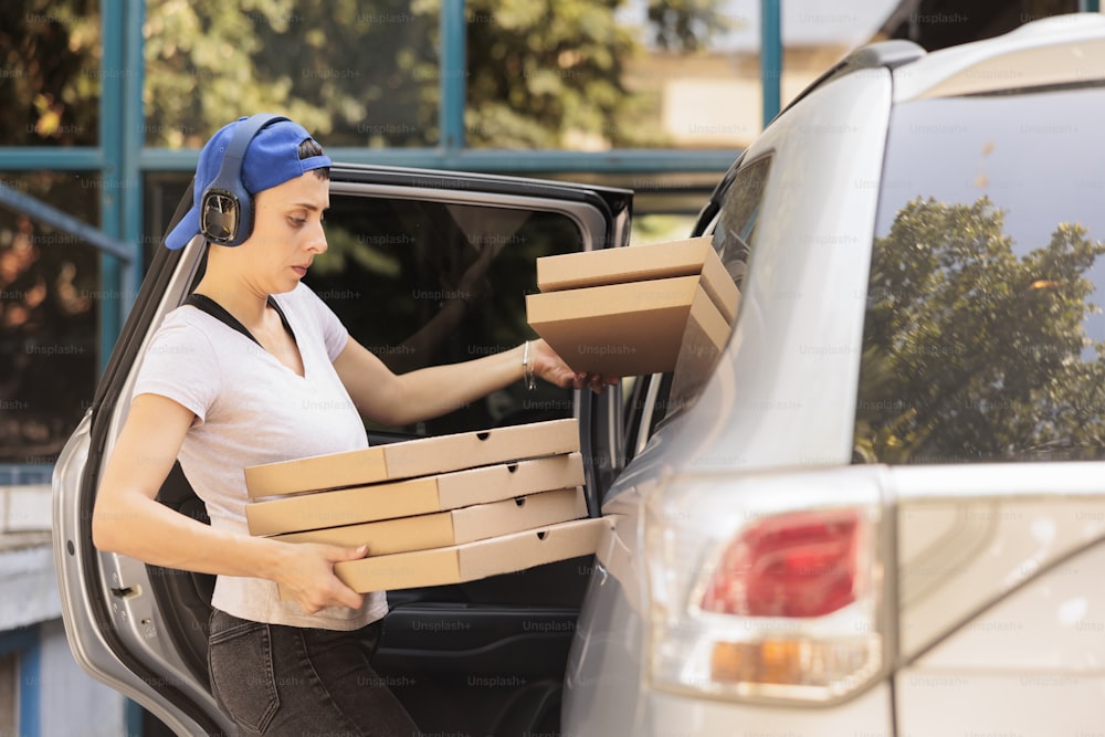 Mulher entregando pizza ao escritório pela vista lateral do carro, jovem correio do restaurante fast food segurando a pilha de caixas. Funcionário caucasiano do serviço de entrega da pizzaria em fones de ouvido carregando almoço