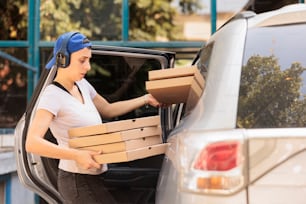 Woman delivering pizza to office by car side view, young fast food restaurant courier holding boxes pile. Pizzeria delivery service caucasian employee in headphones carrying lunch