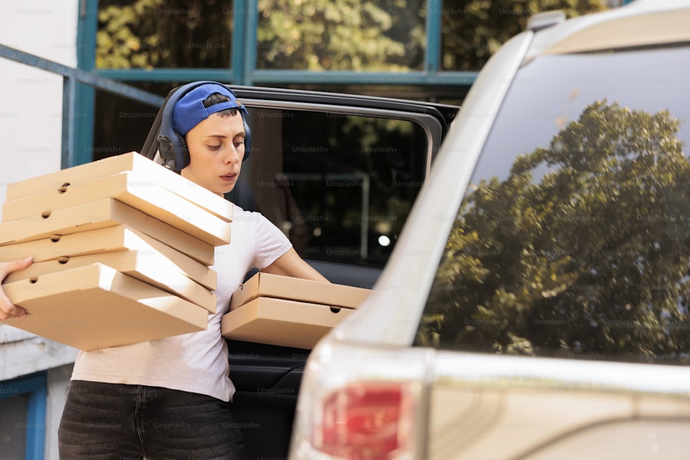 Courier carrying pizza to office by car, young woman holding boxes pile. Pizzeria delivery service employee in headphones carrying lunch, person taking out fast food from vehicle