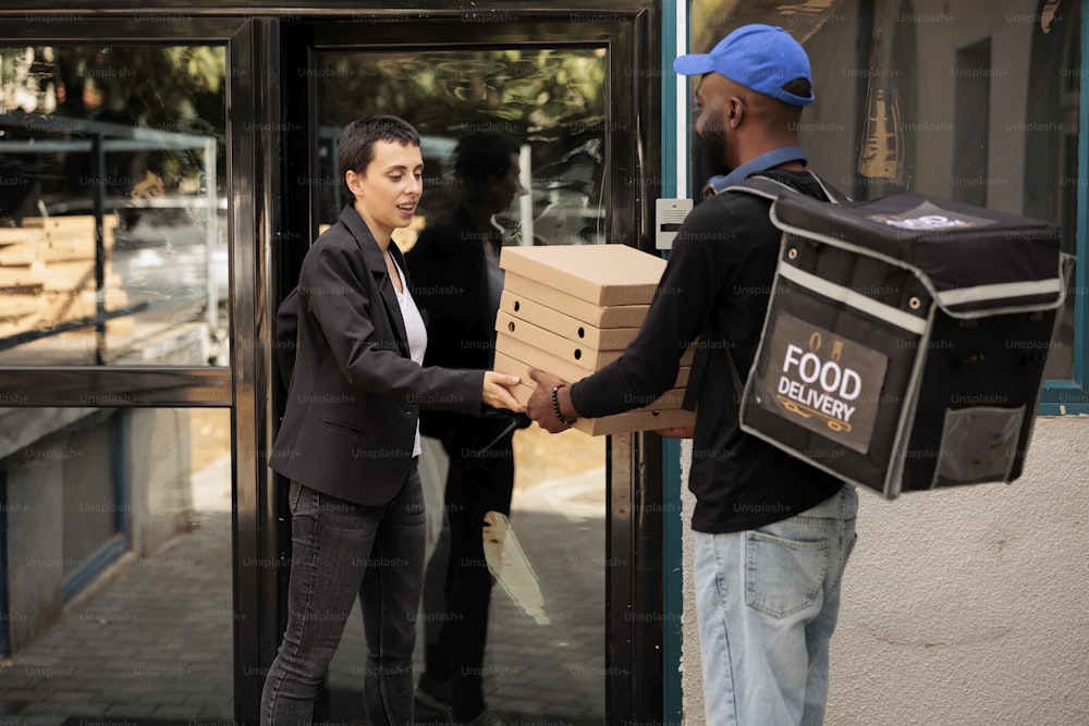Food delivery service courier giving customer pizza order. African american man delivering pizzeria takeaway meal, company employee taking fastfood packages stack near office building