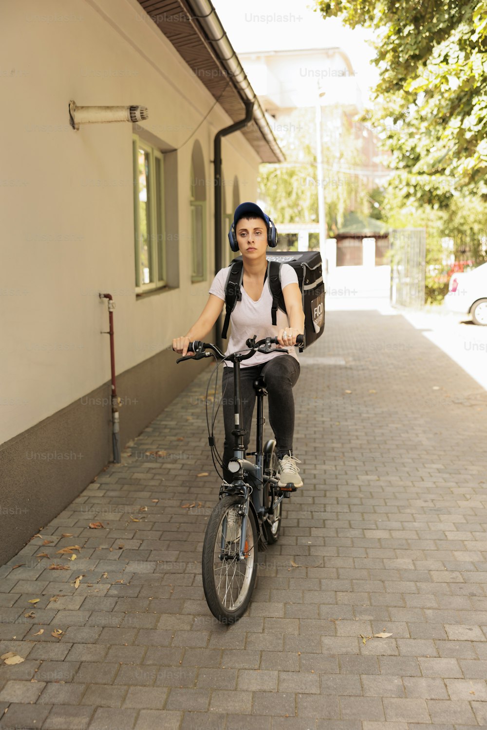 Takeaway food courier in headphones delivering order by bike, front view. Meal delivery service employee riding bicycle with customer order in backpack portrait, woman delivering lunch