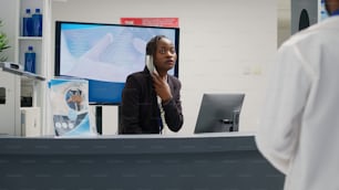 African american woman answering landline phone call at reception counter, working in hospital to give support to patients. Young adult uing telephone with cord at health center front desk.