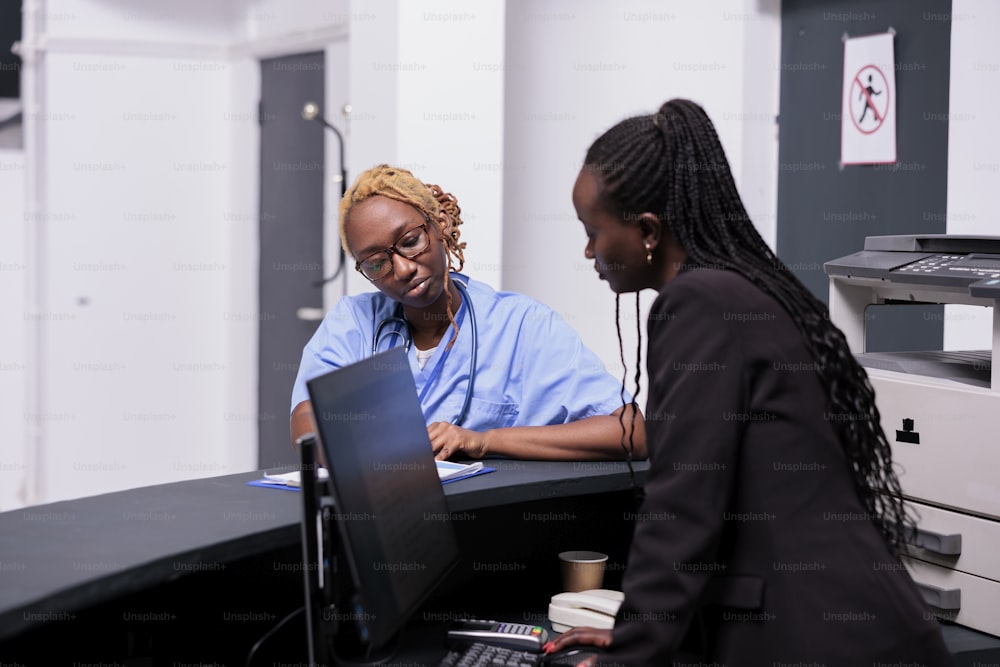 African american medical staff analyzing checkup report with patient insurance support on papers. Nurse and receptionist working on checkup visit appointments at hospital reception desk, healthcare.