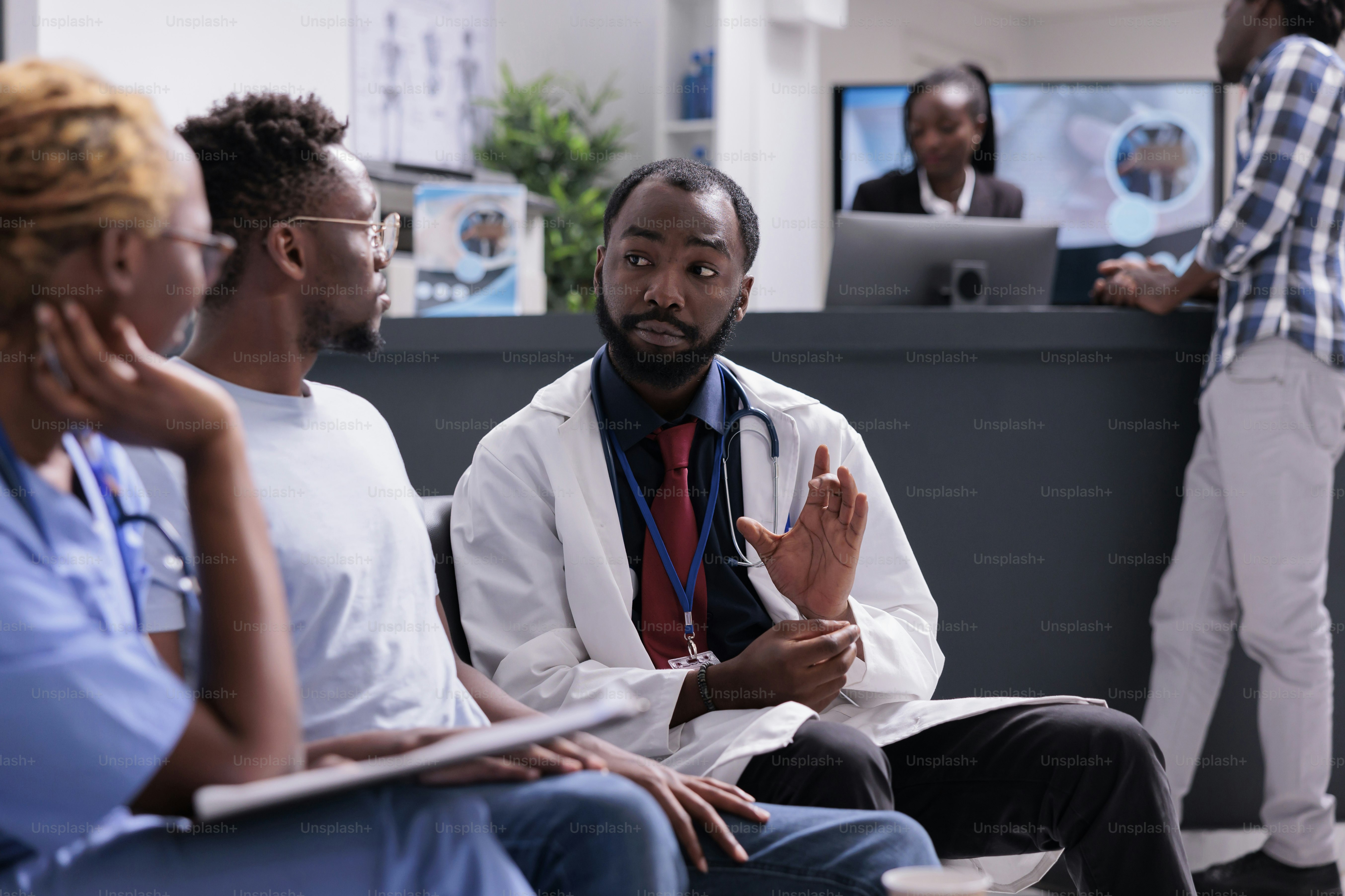 African american medical team consulting man sitting in waiting room at facility reception lobby. Doctor and assistant talking to patient about disease diagnosis and treatment, healthcare support.