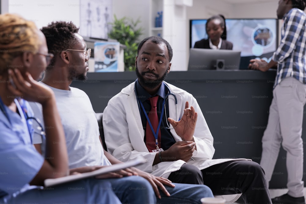 Equipe médica afro-americana consultando homem sentado na sala de espera no saguão de recepção da instalação. Médico e assistente conversando com o paciente sobre diagnóstico e tratamento da doença, suporte à saúde.