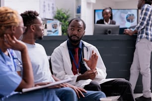 African american medical team consulting man sitting in waiting room at facility reception lobby. Doctor and assistant talking to patient about disease diagnosis and treatment, healthcare support.