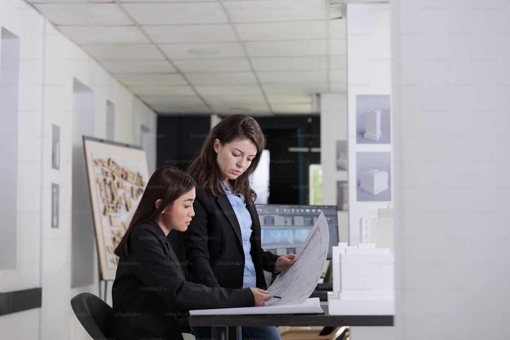 Architects holding blueprint, discussing construction project sketch, working with drafts in coworking space. Employees in architecture office at workplace desk, engineers looking at plan layout