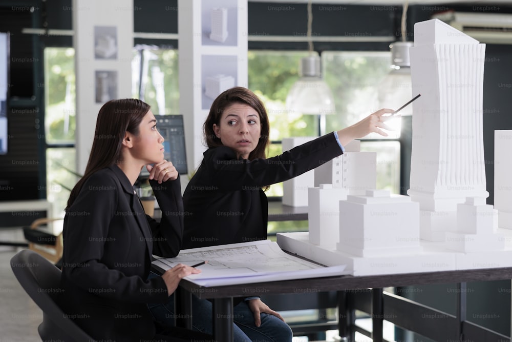 Women working with project draft, discussing building 3d printed model in architecture office. Architect pointing with pencil at architectural prototype in coworking space, construction industry.