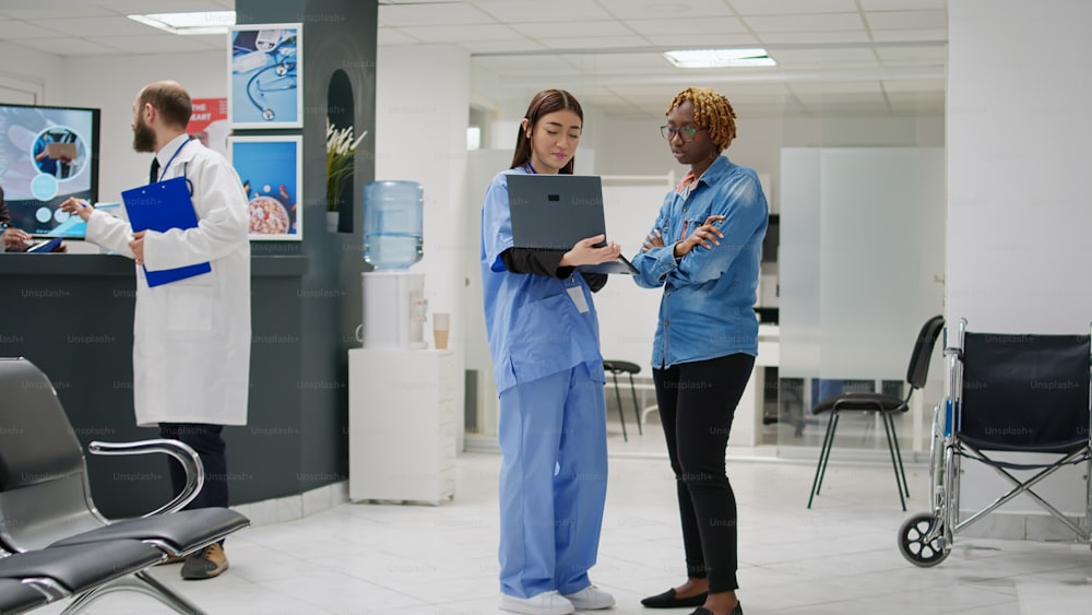 Paciente afroamericano discutiendo con una enfermera sobre la enfermedad y el tratamiento de recuperación, usando una computadora portátil y hablando en el vestíbulo de la sala de espera. Personas charlando en el área de recepción del hospital.