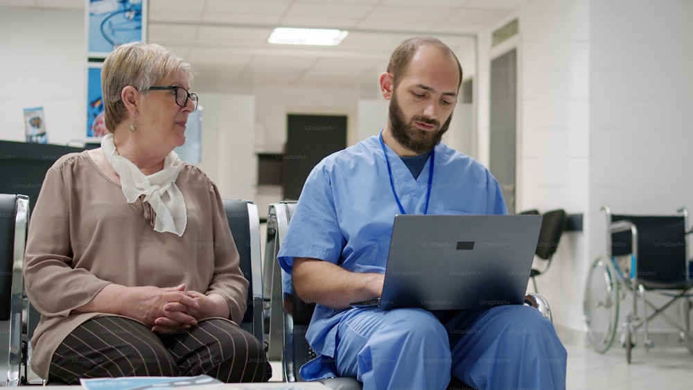 Seniorin und Arzthelferin schaut auf den Laptop in der Empfangshalle des Krankenhauses und spricht über die Diagnose und Behandlung von Krankheiten. Patient und Krankenschwester unterhalten sich über die Genesung im Wartebereich.
