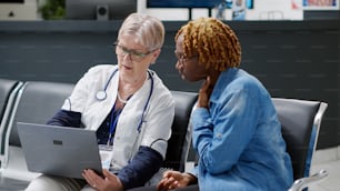 Senior physician showing disease diagnosis to young woman on laptop, explaining treatment at healthcare consultation in medical center lobby. Doing checkup appointment at clinic.