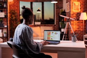 Female analyst looking at data with company research results on laptop screen, back view. African american remote corporate worker analyzing diagrams and charts on computer in modern office in evening