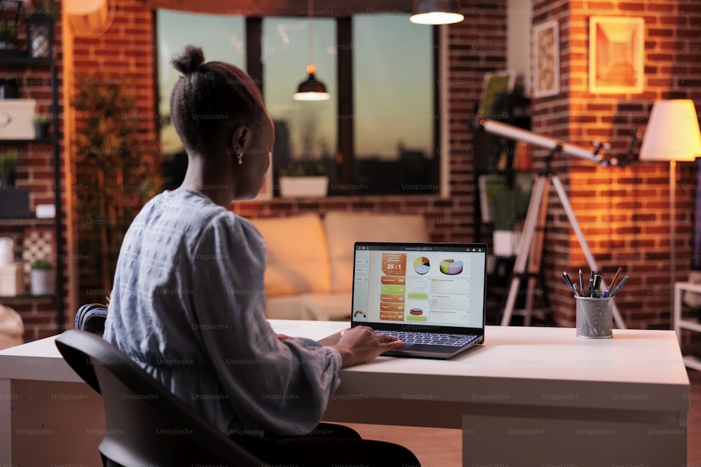 Analista femenina mirando datos con los resultados de la investigación de la compañía en la pantalla de la computadora portátil, vista posterior. Trabajador corporativo remoto afroamericano analizando diagramas y gráficos en la computadora en una oficina moderna por la noche