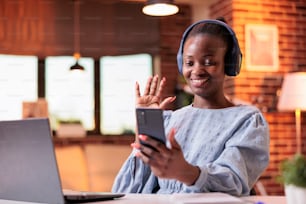 African american woman having online meeting with colleagues on smartphone in modern home office. Smiling female remote worker holding mobile phone and chatting on videconference