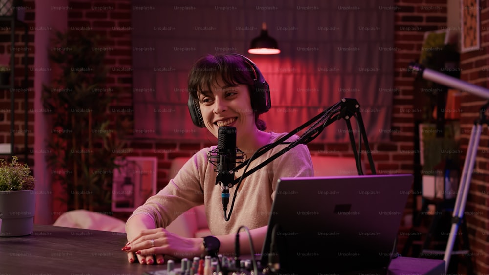 Portrait of woman broadcasting internet radio entertainment show while recording vlog with digital video camera in home podcast studio. Talk show host with headphones smiling while streaming content.