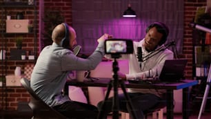 Static tripod shot of internet radio host recording podcast using digital video camera greeting guest in home studio. African american podcaster doing fist bump in interview with famous influencer.