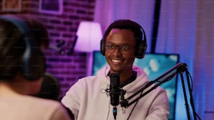 ホームレコーディングスタジオでインフルエンサーとの興味深い会話を聞いている笑顔のアフリカ系アメリカ人オンラインラジオホストのポートレート。ポッドキャスターは、マイクを使用してゲストと一緒にライブ放送します。