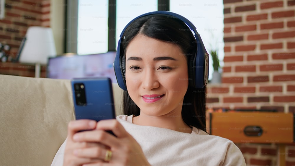 Happy young adult person chatting with friend on modern mobile phone while wearing headphones. Joyful woman sending messages on smartphone while sitting on couch at home.
