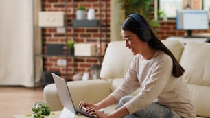 Happy woman wearing headphones while listening music and working from home. Joyful young adult person doing remote work on modern laptop while sitting on couch in apartment.