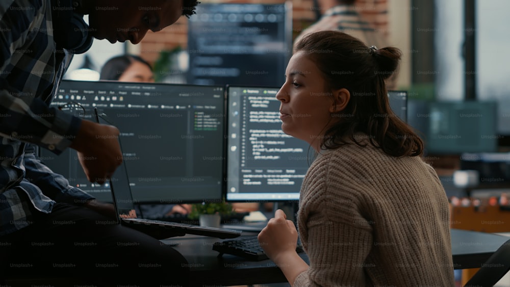 Junior programer sitting down on desk showing laptop with source code to senior dev asking for opinion. Software developer writing algorithm interrupted by colleague wanting help with fixing errors.
