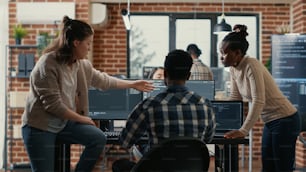 System engineer sitting on desk discussing with mixed team of coders about ai innovation in front of multiple screens compiling code. Programmers doing teamwork looking at machine learning algorithms.