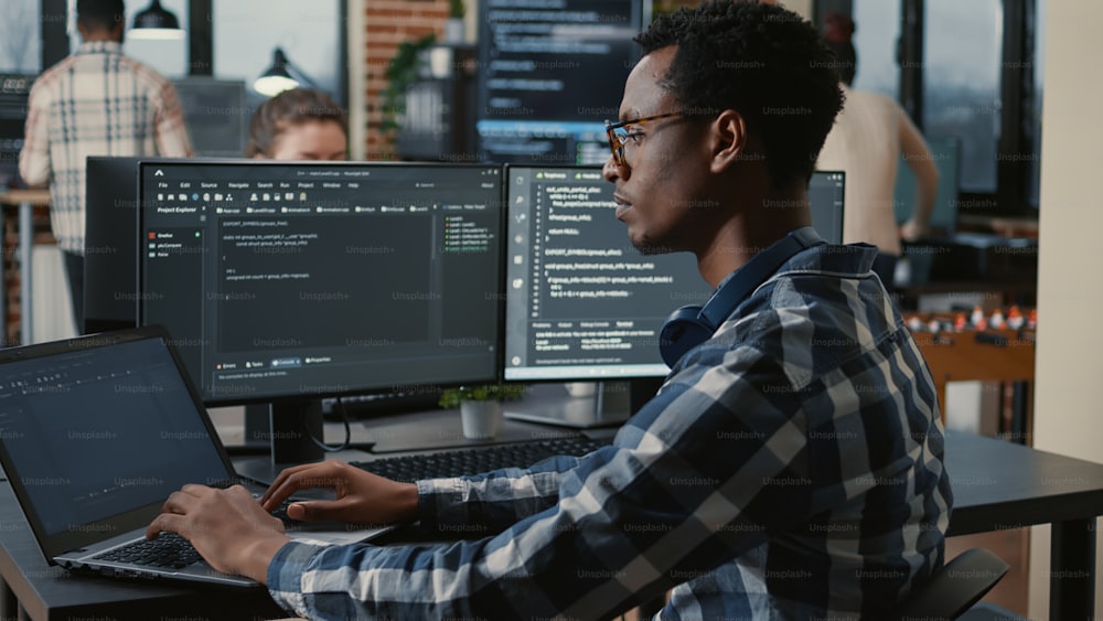 Focused programer fixing glasses while typing on laptop sitting at desk with multiple screens parsing code. Coder working on user interface with team of programmers coding big data in the background.