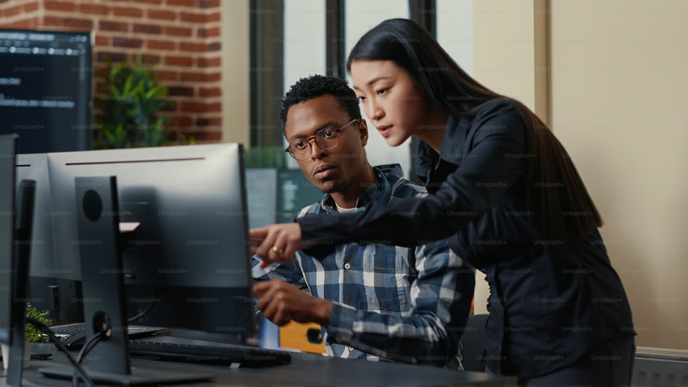 Dos desarrolladores de software que vienen al escritorio y se sientan sosteniendo una computadora portátil con una interfaz de codificación y apuntando a la pantalla de la computadora. Equipo de programadores discutiendo algoritmos mirando monitores con código.